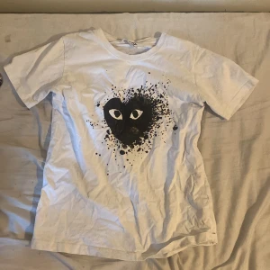 Vit t-shirt med svart hjärta från Comme des Garçons Play - Vit t-shirt från Comme des Garçons Play med ett stort svart hjärta och ögon på bröstet. T-shirten har rund halsringning och korta ärmar. Perfekt för dig som gillar streetwear och vill sticka ut med en ikonisk design.storlek s/m