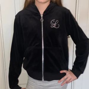Svart velour hoodie med dragkedja och strassdetalj - Säljer en svart hoodie i mjuk velour med dragkedja framtill och siden innanför luvan. På bröstet finns en snygg strassdetalj med bokstäver. Perfekt för en chill och trendig look.