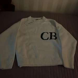 Vit stickad tröja från Cole Buxton - Vit stickad tröja från Cole Buxton med svarta bokstäver 'CB' broderat på bröstet. Tröjan har rund halsringning och ribbade muddar vid ärmslut och nederkant. Perfekt för en stilren och enkel look.