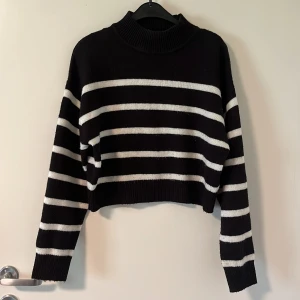 Svartvit randig croppad stickad tröja - Säljer en svart stickad tröja med vita horisontella ränder. Tröjan är croppad, har lång ärm och en hög ribbad krage. Perfekt för dig som gillar en trendig och avslappnad stil.