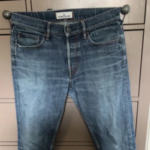 Blå jeans från Stone Island - Säljer ett par klassiska blå jeans från Stone Island med snygg tvätt och ikonisk patch på bakfickan. Jeansen har fem fickor, knappgylf och raka ben. Perfekta för dig som gillar streetwear och vill ha något stilrent men ändå unikt.