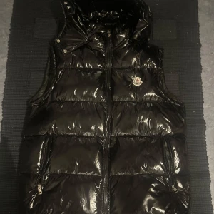 Svart dunväst från Moncler med huva - Säljer en svart, glansig dunväst från Moncler med huva och dragkedja framtill. Västen har två fickor med dragkedja och Moncler-logga på bröstet. Perfekt för lager-på-lager och stilren look.