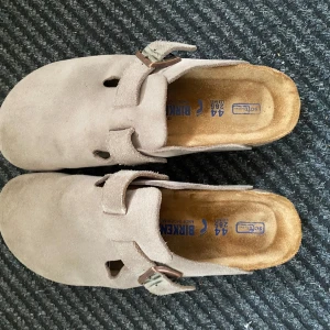Birkenstock Boston - Säljer ett par klassiska Birkenstock sandaler i beige mocka med justerbart spänne och täckt tå. Skorna har en mjuk innersula och platt sula för extra komfort. Perfekta för avslappnade dagar och enkel stil.