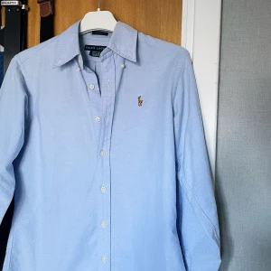Ljusblå skjorta från Ralph Lauren - Säljer en klassisk ljusblå skjorta från Ralph Lauren med broderad logga på bröstet. Skjortan har lång ärm, knappar framtill och är tillverkad i 100% bomull. Perfekt för en stilren och tidlös look. Även perfekt nu till sommaren☀️🏄🌊