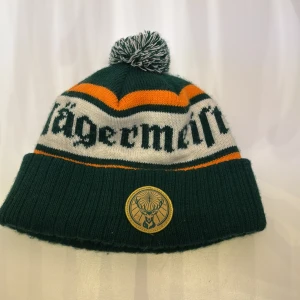 Grön Jägermeister mössa med tofs - Mörkgrön stickad mössa med vit och orange rand, tofs på toppen och Jägermeister-logga framtill. Texten 'Jägermeister' i svart på vit bakgrund runt mössan. Köptes i vintras för 500Kr, använd i bara några dagar.