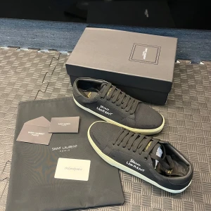 Svarta sneakers från Saint Laurent - Säljer ett par svarta Saint Laurent sneakers med vit sula och broderad logga på sidan. Skorna har snörning och är tillverkade i canvas med detaljer i skinn. Perfekta för en stilren och avslappnad look.