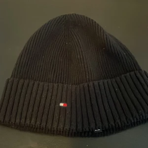 Svart ribbad mössa från Tommy Hilfiger - Svart ribbstickad mössa från Tommy Hilfiger med liten broderad logotyp framtill. Klassisk modell som sitter skönt på huvudet och passar perfekt till kyliga dagar. Hämtas i Västerhaninge tar endast kontanter.