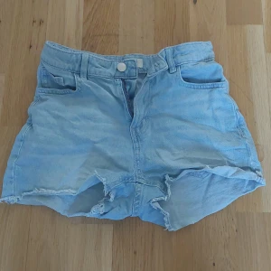 Ljusblå jeansshorts med fransar - Säljer ett par ljusblå jeansshorts med råa franskantade benslut och klassisk femficksdesign. Shortsen har knapp och dragkedja framtill samt bälteshällor i midjan. Perfekta för varma dagar!