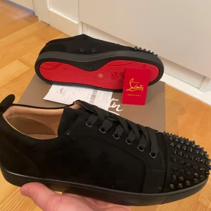 Svarta Christian Louboutin med nitar - Säljer ett par svarta sneakers från Christian Louboutin med ikonisk röd sula och svarta nitar framtill. Perfekta för dig som vill sticka ut med en lyxig och edgy stil.
