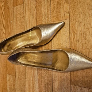 Guldiga pumps från Aldo Castagna - Säljer ett par eleganta pumps i guldigt skinn från Aldo Castagna. Skorna har spetsig tå och klack, vilket ger en stilren och lyxig känsla. Perfekta för dig som vill sticka ut med en glansig finish och klassisk design. Används en gång