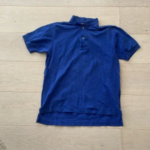 Blå pikétröja från GANT - Säljer en klassisk blå pikétröja från GANT i storlek M. Tröjan har korta ärmar, krage och knäppning med två knappar framtill. Perfekt för en avslappnad och stilren look.