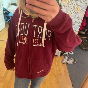 Vinröd hoodie med dragkedja från FOX Originals - Säljer en vinröd hoodie från FOX Originals med vit dragkedja och snören. På bröstet står det 'PORTUGAL' i stora bokstäver och det finns en patch på ena ärmen. Hoodien har två fickor framtill och mjukt material.seljer eftersom den är för stor har i vanliga fall S, tröjan är stor i stoleken.Priset kan diskuteras.
