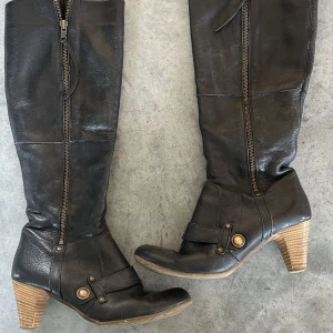 Svarta höga stövlar med dragkedja och klack - Säljer ett par svarta höga stövlar i skinn med dekorativa knappar och dragkedja längs hela skaftet. Snygg klack i trälook och rund tå. Perfekta för att lyfta din outfit under kallare månader.