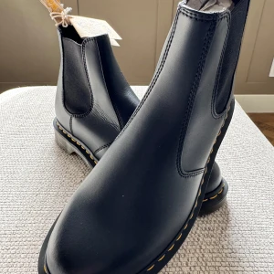 Svarta chelsea boots från Dr. Martens - Säljer ett par aldrig använda klassiska svarta chelsea boots från Dr. Martens med gul söm runt sulan. Skorna är i skinn och har elastiska paneler på sidorna för enkel påtagning. Perfekta för dig som vill ha en tidlös och stilren look.