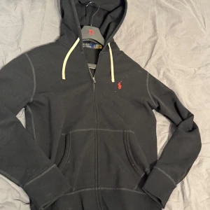 Svart hoodie från Polo Ralph Lauren - Svart hoodie från Polo Ralph Lauren med dragkedja och vit dragsko i huvan. Klassisk röd logga broderad på bröstet och två fickor framtill. Perfekt för en avslappnad stil. Den är knappast använd och är i väldigt bra skick det är storlek S och fraktas direkt vid köp