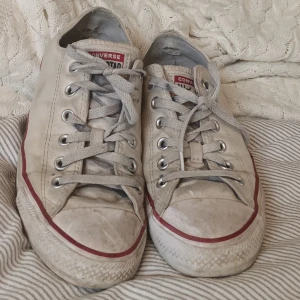 Vita Converse All Star sneakers - Klassiska vita Converse All Star sneakers med lågt skaft och röd rand längs sulan. Skorna har snörning och rund tå, samt den ikoniska loggan baktill. Perfekta för en avslappnad och tidlös stil.