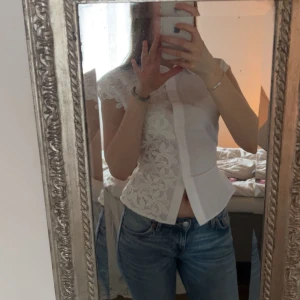 Vit blus med spetsdetaljer och rosett i ryggen - Säljer en vit blus med vackra spetsdetaljer framtill och korta ärmar. Blusen har en feminin passform och knäppning framtill. Perfekt att styla med jeans för en fräsch look.