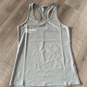 Grå linnetopp från Vitamin Well - Säljer en grå sportig linnetopp från Vitamin Well med brottarrygg. Perfekt för träning och har en enkel, stilren design med logga framtill. Tillverkad i ett mjukt och stretchigt syntetmaterial.
