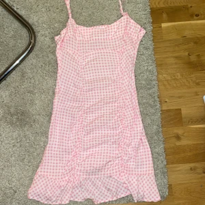 Rosa och vit rutig klänning från Divided - Supersöt rosa och vit rutig klänning från Divided med smala axelband och figurnära passform. Perfekt för sommardagar och har en lätt volang nertill. Klänningen är i ett mjukt och tunt material som känns svalt.
