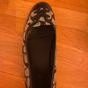 Svart-beige ballerinaskor med snörning från Coach - Säljer ett par ballerinaskor från Coach i svart och beige med tydligt logomönster. Skorna har svarta snören framtill och svarta detaljer i glansigt material vid tån. De är i perfekt skick aldrig använda av mig då de var för stora. 