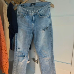 Ljusblå slitna jeans med bootcut - Säljer ett par ljusblå jeans med slitningar på låret och knät. Jeansen har bootcut-modell och råa, fransiga benslut. Klassisk femficksdesign och normal passform. Perfekta för en avslappnad och trendig look.