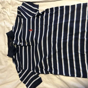 Randig pikétröja från Ralph Lauren - Säljer en stilren randig pikétröja från Ralph Lauren i marinblått och vitt. Tröjan har en klassisk krage med knappar och det ikoniska röda broderade logotypen på bröstet. Perfekt för en avslappnad men ändå snygg look.