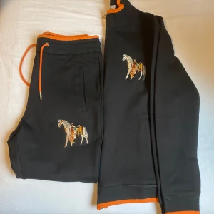 Svart hoodie med hästbroderi - Säljer en svart hoodie med dragkedja och broderat hästmotiv på bröstet. Hoodien har orange detaljer vid krage och ärmslut, vilket ger en snygg kontrast. Perfekt för en avslappnad stil. Passar som S/M
