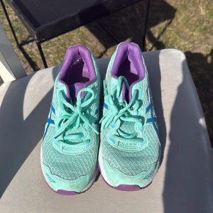 Mintgröna träningsskor från Asics - Säljer ett par fräscha mintgröna träningsskor från Asics med lila detaljer. Skorna har snörning och en bekväm passform, perfekt för löpning eller gymmet. De har en rund tå och en stabil sula för bra grepp.