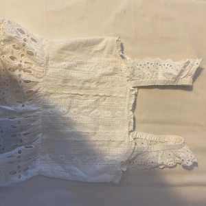 Vit spetsprydd topp - Charmig vit topp med vackra spetsdetaljer och broderier. Toppen har en smockad del för bättre passform och ärmlös design med volangdetaljer. Perfekt för sommarens varma dagar.