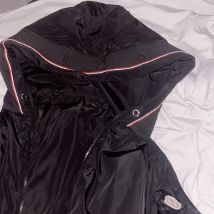 Svart jacka från Moncler - Snygg svart jacka från Moncler med huva och dragkedja. Jackan har en stilren design med Moncler-logga på ärmen och detaljer i rött och vitt. Perfekt för en cool och avslappnad stil. Lite stor i storleken men passar endå🩷
