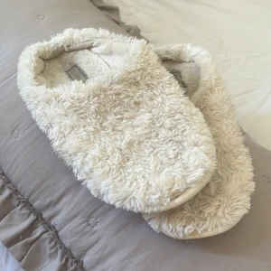 Vita fluffiga tofflor - Mysiga vita tofflor med fluffigt material som håller fötterna varma. Perfekta för att glida runt hemma under kyliga dagar. De har en halkfri sula för extra säkerhet.