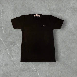 Svart t-shirt från Comme des Garçons - Säljer en svart t-shirt från Comme des Garçons med ett ikoniskt hjärta med ögon broderat på bröstet. T-shirten är kortärmad och gjord i mjuk bomull. Perfekt för en stilren look.
