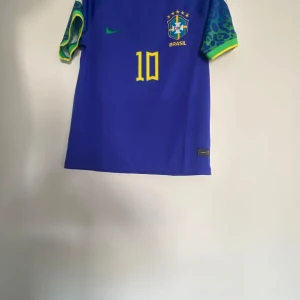 Blå Brasilien fotbollströja - Säljer en blå Brasilien fotbollströja från Nike med nummer 10 och Neymar Jr på ryggen. Tröjan har korta ärmar med grönt mönster och gula detaljer. Perfekt för fotbollsfans! Storlek M passar L