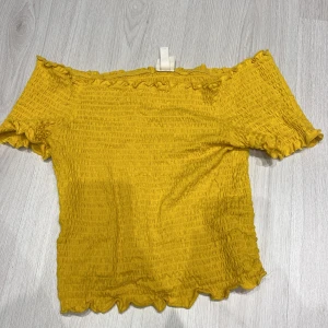 Gul offshoulder topp från H&M - Säljer en söt gul offshoulder topp från H&M. Toppen har en smockad design med volangdetaljer vid ärmslut och nederkant. Perfekt för en solig dag!