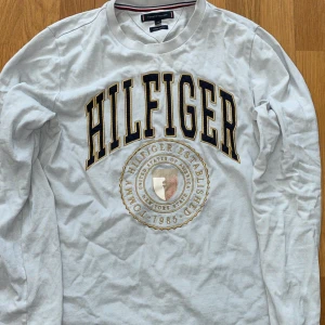 Tommy Hilfiger - Säljer en vit långärmad tröja från Tommy Hilfiger med stort broderat märke på framsidan. Tröjan har en rund halsringning och är perfekt för en avslappnad stil. Passar bra till jeans eller chinos.