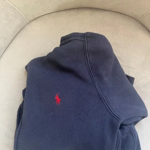 Marin blå hoodie från Ralph Lauren - Säljer en stilren marin blå hoodie från Ralph Lauren med det klassiska röda logotypbroderiet på bröstet. Tröjan har en dragkedja framtill och är perfekt för en avslappnad look. Ord pris 1199kr