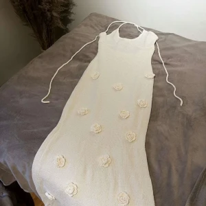 Beige stickad klänning med blommor - Charmig beige stickad klänning med dekorativa blommor. Klänningen har smala axelband och en figurnära passform. Perfekt för en stilren och romantisk look.