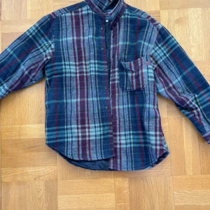 Rutig flanellskjorta från Exit - Säljer en snygg rutig flanellskjorta från Exit i blått, rött och grönt. Skjortan har långa ärmar och en bröstficka med knapp. Perfekt för en avslappnad stil. Tillverkad i Italien.