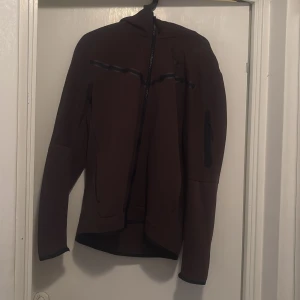 Nike tech brun - Säljer en brun kofta från Nike med dragkedja och huva. Koftan har en stilren design med långa ärmar och diskreta detaljer. Perfekt för en sportig look eller en avslappnad dag. Koftan är i storlek L och använd ett par gånger hemma. Aldrig utomhus 