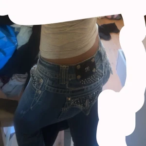 Blå jeans med strassdetaljer från Miss Me - Snygga blå jeans från Miss Me med glittrande strassdetaljer på bakfickorna och framsidan. Jeansen har en låg midja och en utvsvängd passform. Mått: midja: 37,5cm innerbenslängden: 84cm
