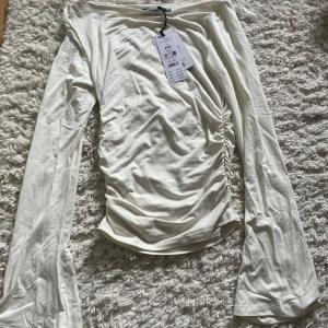 Vit långärmad topp från Gina  - Säljer en stilren vit långärmad topp från Gina med rynkade detaljer på sidan. Toppen har en elegant passform och är perfekt för både vardag och fest. Materialet är mjukt och bekvämt. HELT NY MED PRISLAPP