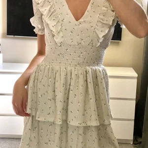 Vit blommig klänning med volangdetaljer - Charmig vit klänning med små blommor och volangdetaljer. Den har en v-ringad hals och korta ärmar. 