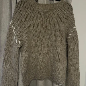 Beige stickad tröja med snörning - Säljer en mysig beige stickad tröja med unik snörning längs ärmarna. Perfekt för kyliga dagar och ger en stilfull touch till din outfit. Tröjan har en avslappnad passform och rund halsringning.