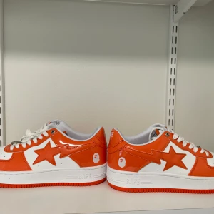 Orange och vita sneakers från BAPE - Snygga BAPE sneakers i en fräsch orange och vit färgkombination. Skorna har en glansig finish med det ikoniska BAPE-stjärnmärket på sidan och en metallisk detalj vid snörningen. Perfekta för att sticka ut i mängden!