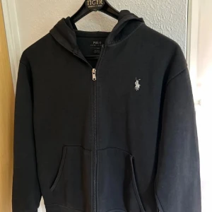 Svart hoodie från Polo Ralph Lauren - Säljer en stilren svart hoodie från Polo Ralph Lauren med dragkedja och det klassiska logotypbroderiet på bröstet. Tröjan har långa ärmar och en praktisk huva. Perfekt för en avslappnad look. Storlek 170 typ S.