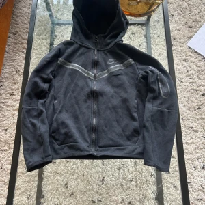 Svart Nike tech hoodie - Säljer en svart hoodie från Nike med dragkedja och huva. Jackan har långa ärmar och ett diskret Nike-logotyp på bröstet. Perfekt för en sportig look eller avslappnade dagar.