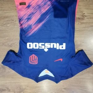 Blå fotbollströja från Nike - Säljer en blå fotbollströja från Nike med korta ärmar och ett coolt mönster i rosa och blått. Tröjan har en vit logga på framsidan och en orange Nike-swoosh. Perfekt för fotbollsfans!