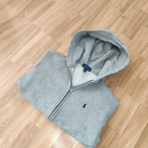 Grå hoodie från Ralph Lauren - Säljer en stilren grå hoodie från Ralph Lauren med dragkedja och en liten logga på bröstet. Perfekt för en avslappnad look. Den har en mjuk insida och en praktisk huva.