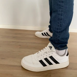 Vita sneakers från Adidas - Snygga vita sneakers från Adidas med klassiska svarta ränder på sidorna. Skorna har en rund tå och snörning framtill. Perfekta för en avslappnad stil.