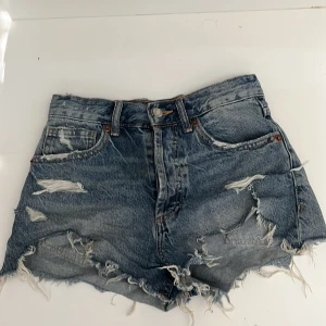Blå jeansshorts från Zara - Säljer ett par coola blå jeansshorts från Zara med slitna detaljer och fransar. Perfekta för en avslappnad stil med hög midja och klassisk femficksdesign. Lite trasig på högra benet